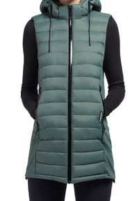 Bezrękawnik w zielonym kolorze, pikowany, z kapturem i czarnym zamkiem, noszony na czarnej bluzce z długim rękawem, ręce w kieszeniach bocznych.