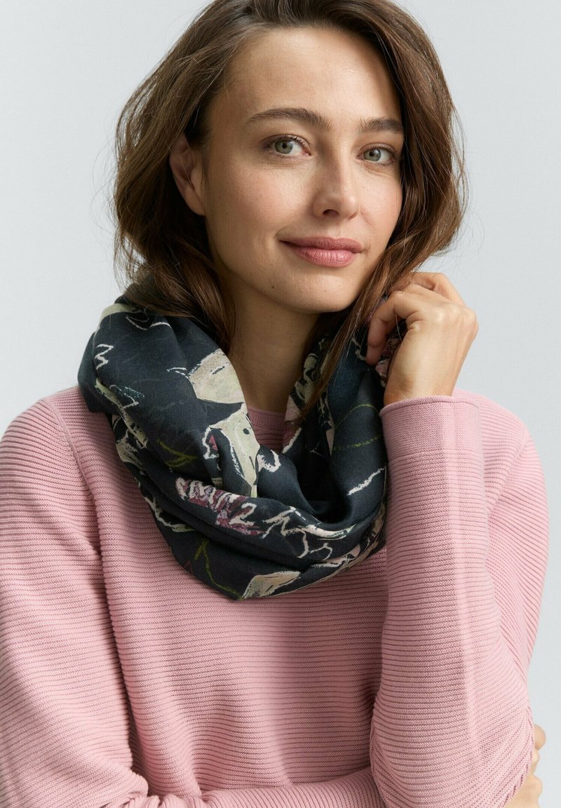 Vrouw met lichtbruin haar die een roze geribbelde trui en een donkere bloemenpatroon sjaal draagt, zacht glimlachend tegen een effen achtergrond.