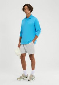 Helles blaues Poloshirt mit langen Ärmeln und texturiertem Muster, graue Shorts, cremefarbene Basecap und beige Turnschuhe mit grünen Akzenten.