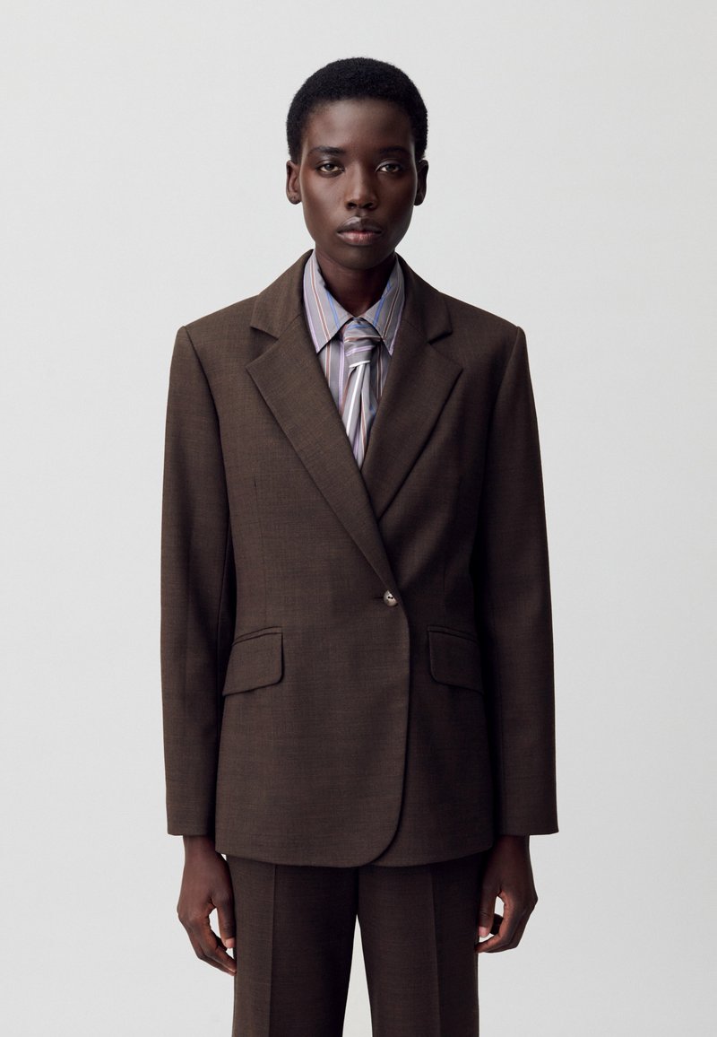 Blazer marrone su misura con due tasche frontali, revers a punta e chiusura con un solo bottone, abbinato a una camicia a righe e pantaloni coordinati.