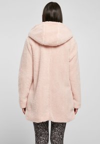 Cappotto in pelliccia sintetica rosa testurizzata con un ampio cappuccio, dotato di spalle scese e vestibilità rilassata, abbinato a leggings neri con fantasia.