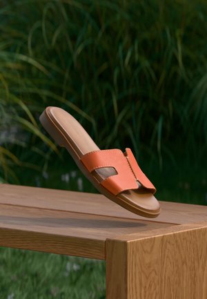 Orange slide sandal with two large cutouts, textured leather upper, flat beige sole, and a smooth finish, displayed on a wooden surface.