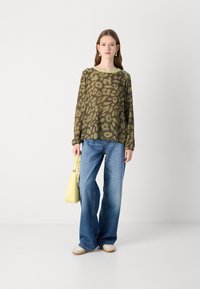 Olijfgroene blouse met een luipaardprint, lange mouwen, ronde halslijn met gele afwerkingsrand, gecombineerd met losse blauwe jeans en een gele handtas.