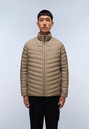 Beige quiltet pufferjakke med høj krave, synlig lynlås og struktureret overflade. Båret over mørke bukser mod en lys baggrund.
