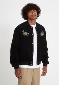 Homme aux cheveux bouclés portant une veste noire boutonnée ornée de scorpions brodés sur la poitrine, par-dessus une chemise blanche et un pantalon beige.