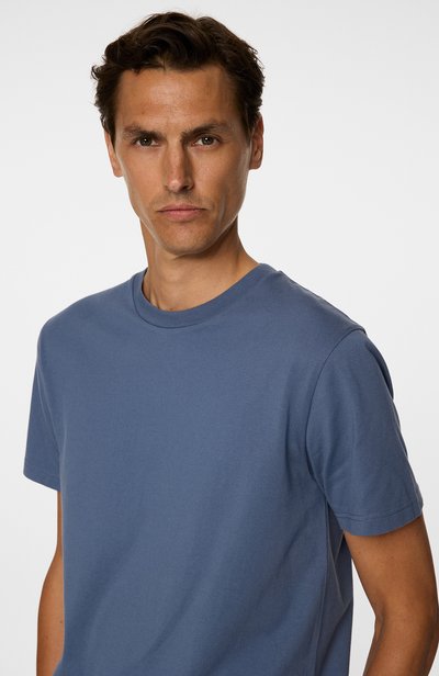 T-shirt en coton bleu avec un col rond et des manches courtes. Le tissu semble lisse avec une texture subtile. Le modèle pose de face.