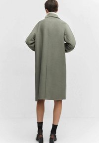 Long manteau vert olive avec un col blanc doux, coupe droite et poches latérales. Le tissu semble texturé ; le design est minimaliste et fonctionnel.