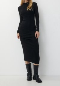 Femme portant une robe midi noire ajustée à manches longues, associée à des bottes en cuir noires mi-mollet, se tenant devant un fond blanc uni.
