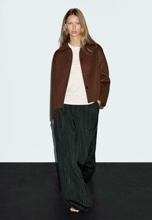 Veste en laine marron avec un col et une fermeture à boutons, associée à un t-shirt léger et un pantalon large à rayures noires. Textures lisses, coupe décontractée.
