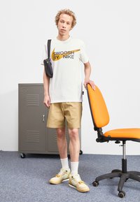 Bílý tričko s hnědým grafickým motivem "SWOOSH BY NIKE", khaki kraťasy, bílé ponožky a žluté tenisky. Černá taška a oranžová kancelářská židle viditelné.