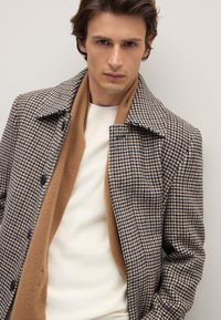 Op maat gemaakte pied-de-poule jas in bruin en marineblauw, met een brede kraag, zwarte knopen en een zachte bruine sjaal over een crème trui.