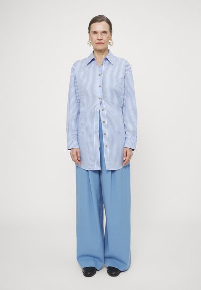 Striped blue button-up shirt with long sleeves and a slightly elongated cut; paired with loose blue trousers and black shoes.