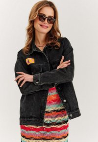 Czarna kurtka dżinsowa z jasną naszywką z logo, ma długie rękawy i kieszenie z przodu. Noszona na kolorowej sukience w wzory.