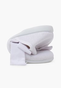 Zapatilla de malla blanca con suela suave, parte superior de felpa y acento de lazo. El diseño es compacto y ligero con una superficie texturizada.