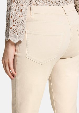 Close-up af en person iført cremefarvede jeans med en synlig baglomme og en beige hæklede langærmet top.