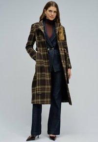 Mujer vestida con un abrigo largo de cuadros marrón y verde sobre una chaqueta oscura, un suéter de cuello alto marrón, jeans de pierna ancha y tacones puntiagudos marrones.