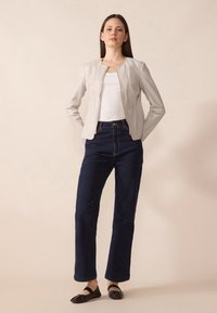 Lys grå skinjakke med rund hals, kombinert med høyt midje mørkeblå denimbukser. Modellen har på seg svarte flate sko.
