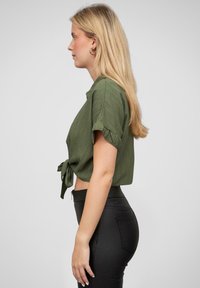 Cămașă scurtă, cu mânecă scurtă, verde măsliniu, cu un nod în față, realizată dintr-un material texturat. Asortată cu pantaloni strâmți negri, cu talie înaltă.