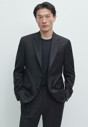 Uomo con abito nero e camicia nera in piedi con le mani nelle tasche contro uno sfondo chiaro e uniforme.