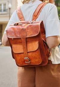 Personne portant une chemise blanche et un pantalon marron porte un grand sac à dos en cuir marron avec une poche avant à boucle dans une rue de la ville.
