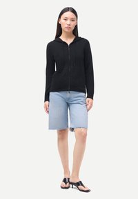 Svart zip-up-hoodie med texturerad yta, utrustad med huva och dragsko, kombinerad med ljusblåa jeansshorts och svarta sandaler.