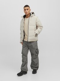 Hellgraue Pufferjacke mit Kapuze, großen Taschen und schwarzen Akzenten, kombiniert mit weiten dunkelgrauen Cargo-Hosen und schwarzen Stiefeln.