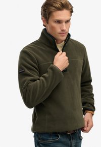 Olive zielony polarowy sweter z półzamkiem, z kontrastującą ciemnoniebieską podszewką i naszywką z logo na rękawie. Gładka faktura.