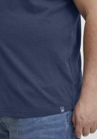 Tamsiai mėlyna medvilninė marškinėliai su apvalia iškirpte, trumpomis rankovėmis ir šoniniu etiketo akcentu. Turi lygią tekstūrą ir klasikinį siluetą.