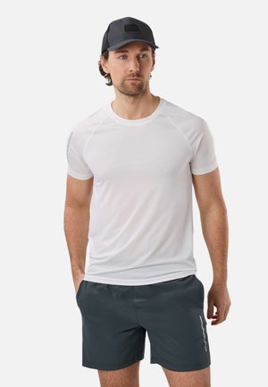 Männliches Model trägt ein weißes, kurzärmeliges Sportshirt, dunkelgraue Shorts und eine schwarze Baseballkappe und steht mit den Händen in den Taschen.
