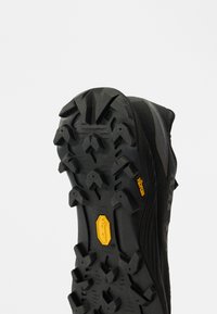 Suola di scarpe da trail running nere con battistrada profondo e aggressivo, e logo Vibram giallo. Design in gomma testurizzata per una maggiore aderenza e durabilità.