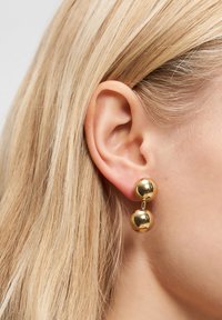 Goldene Ohrringe aus Metall mit zwei sphärischen Komponenten, die durch einen dünnen Stift verbunden sind, und einer glatten, reflektierenden Oberfläche.