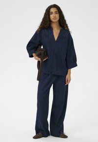 Sieviete ar gariem viļņotiem matiem, tērpusies brīvā tumši zila džinsu blūzē un biksēs, tur brūnu somu, stāvot pretī vienkāršai gaišai fona.