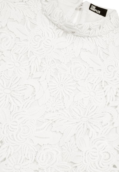 Tissu en dentelle blanche avec des motifs floraux complexes et un petit col, arborant une étiquette noire avec le texte "THE KOOPLES".