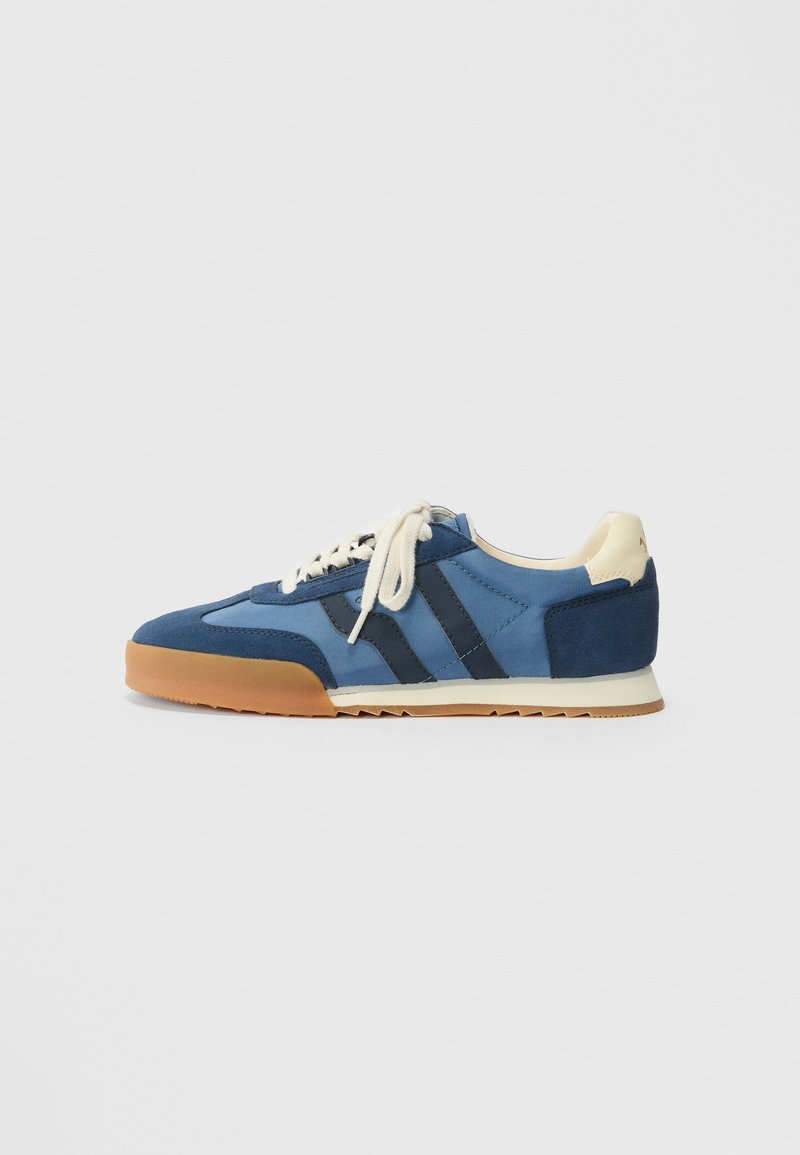 Blå sneakers laget av semsket skinn og stoff, med marineblå striper, beige lisser og en gummisåle med zigzag mønster for godt grep.