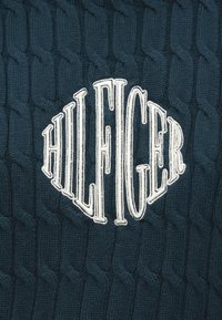 Textil tricotat de culoare albastru închis, având un model de cablu ridicat, cu un logo brodat în alb "HILFIGER" în formă rotundă, situat în centru.