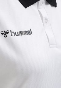Gros plan d'un polo blanc avec col noir et logo « hummel » noir sur la poitrine, montrant deux boutons sous le col.