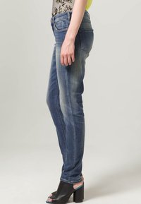 Jeans in denim blu slim-fit con texture sbiadita e leggere pieghe, abbinati a tacchi aperti neri con accenti blu.