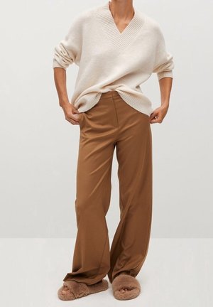 Person iført en cremefarvet V-hals sweater, brune bukser med vidde i benene og brune, pelsede åbne tå-sandaler, stående mod en ensfarvet baggrund.