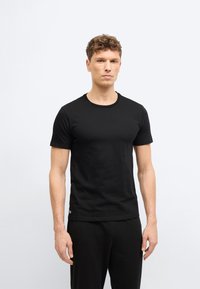 Άνδρας με κοντά, σγουρά μαλλιά, φορώντας εφαρμοστό μαύρο κοντομάνικο μπλουζάκι και μαύρο παντελόνι, στέκεται μπροστά σε απλό φωτεινό φόντο.