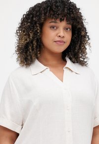 Short-sleeve, button-up shirt in a light beige fabric. Features a collared neckline and a textured surface with subtle stitching detail.