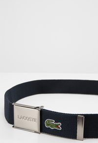 Navy woven belt with a metal clasp featuring a silver-finished buckle and Lacoste logo. Includes a green embroidered crocodile logo detail.