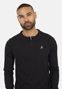 Zwarte lange mouwen polo met een rits, ribgebreide kraag en een klein geel vogellogo op de linkerkant van de borst. Zachte stoftextuur.