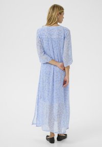 Maxi dress in light blue sheer fabric with intricate floral patterns. Features three-quarter sleeves and a relaxed fit. Black flat shoes.