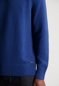 Sininen neulottu villapaita, jossa on teksturoitu kuvio, resorit hihansuissa ja helmassa, sekä pieni lappu vasemmassa alareunassa.