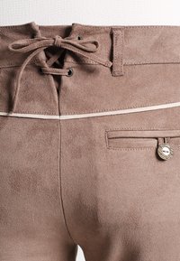 Bruine faux suede broek met een vetersluiting aan de voorkant, witte accentstiksels en een kleine achterzak met een ronde zilveren knop.