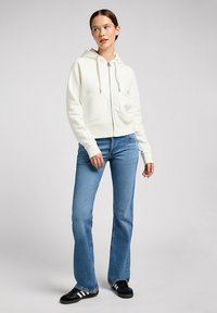 Weißer Zip-Hoodie mit Kapuze und zwei Fronttaschen, kombiniert mit blauen Schlagjeans und schwarzen Sneakern mit weißen Streifen.