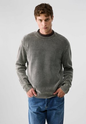 Jeune homme aux cheveux bouclés portant un pull en laine gris et un jean bleu, debout, les mains dans les poches, sur un fond uni.
