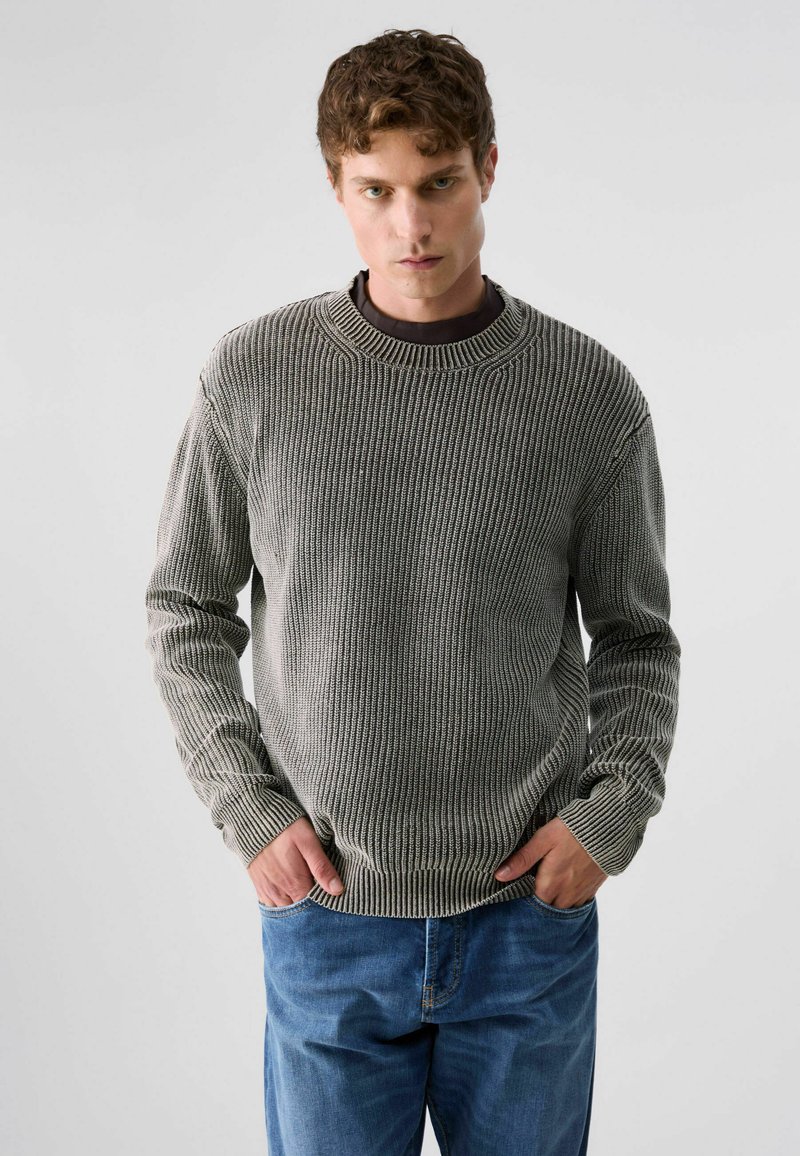 Jeune homme aux cheveux bouclés portant un pull en laine gris et un jean bleu, debout, les mains dans les poches, sur un fond uni.