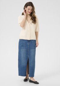 Gilet en tricot beige clair à manches courtes, avec des boutons sur le devant, accompagné d'une jupe en denim longue présentant une fente à l'avant et une teinte bleue. Chaussures plates noires.