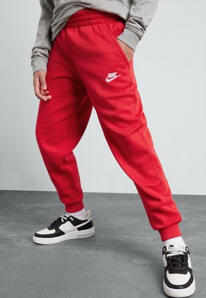 Personne portant un pantalon de survêtement Nike rouge et des baskets Nike noir et blanc, debout sur un sol gris devant un fond uni.